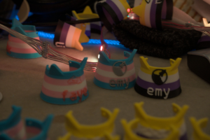 Verschiedene Katzenohren-Mate Tags in Trans und Nicht-Binär Design liegen verteilt auf einem Tisch, mit anderem Krimskrams und einer Lichterkette darauf.