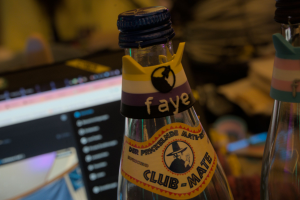 Ein Katzenohren-Mate Tag im Nicht-Binär Design mit meinem Namen und dem vspace.one Logo drauf auf einer Flasche Club-Mate. Im Hintergrund mein offener Laptop und ein Tisch mit viel Krimskrams. Rechts ist eine Flasche, die ein Tag im Trans Design an sich hängen hat zu sehen.