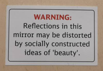 Weißer Sticker mit rotem Text "WARNING:", darunter in Schwarz "Reflections in this mirror may be distorted by socially constructed ideas of 'beauty'.", drum herum ein schwarzer Rahmen mit abgerundeten Ecken. Der Sticker liegt auf einem Holzbrett.